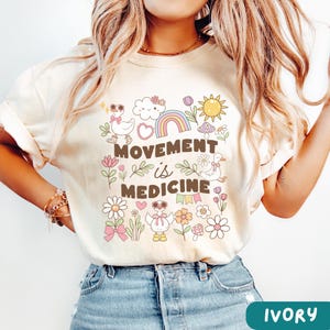 May include: Ivory t-shirt with a graphic design featuring a rainbow, clouds, flowers, mushrooms, and the text "Movement is Medicine".
