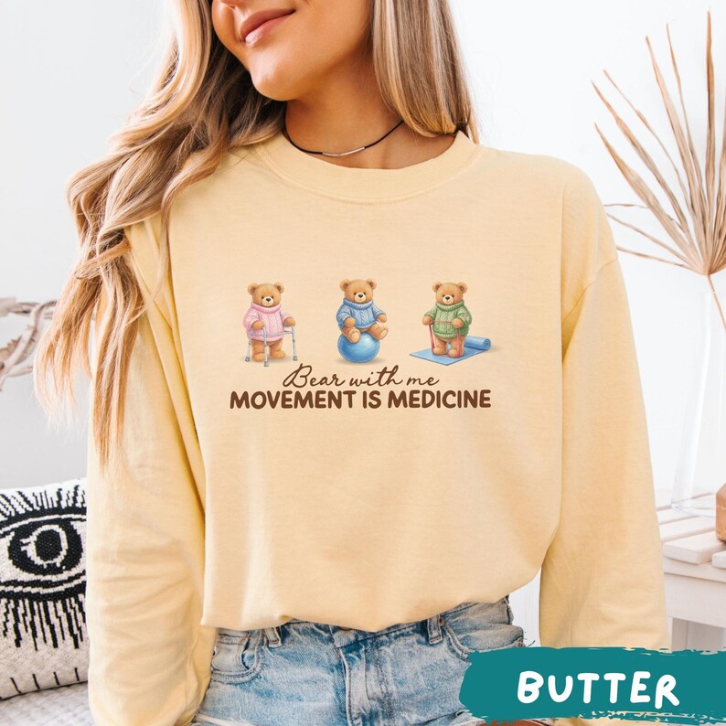 May include: A light yellow sweatshirt with the text "Bear with me, Movement is Medicine." The graphic features three teddy bears: one with a cane, one on a blue exercise ball, and one on a yoga mat. The word "BUTTER" is at the bottom.