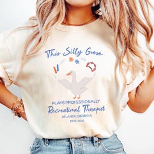 May include: A white t-shirt with a blue and red illustration of a goose juggling a puzzle piece, crayons, a tambourine, and a red and white striped object. The text "This Silly Goose Plays Professionally Recreational Therapist Atlanta, Georgia Estd 2025" is printed below the illustration.