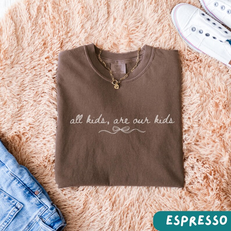 May include: Espresso-colored t-shirt with the text "all kids, are our kids" in a cursive font. A gold chain necklace with a pendant rests on the shirt. The shirt is folded on a fluffy, light brown surface, with a pair of white sneakers and blue jeans visible.