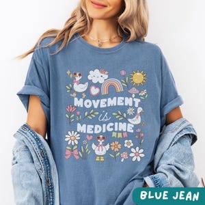 May include: A blue jean t-shirt with a graphic design featuring a rainbow, clouds, flowers, and the text "Movement is Medicine".