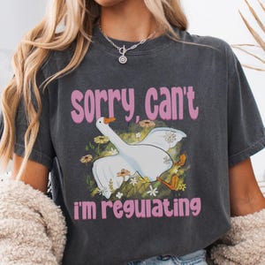 Peut inclure: T-shirt gris foncé avec l'inscription rose "sorry, can't" au-dessus d'une illustration de dessin animé d'une oie. L'oie est blanche avec des pieds oranges et un bec jaune, entourée de fleurs, et le texte "I'm regulating" est en dessous.