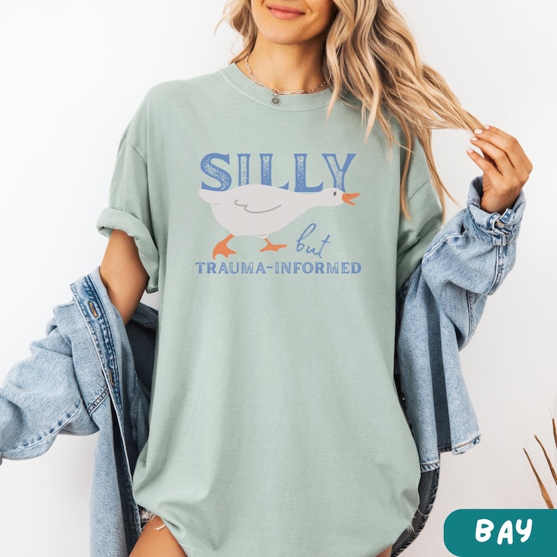 May include: A light green t-shirt with the words "SILLY but TRAUMA-INFORMED" in blue text. A white cartoon goose with orange feet and beak is featured in the center. The shirt has short sleeves and a relaxed fit.