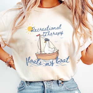 May include: Cream-colored t-shirt featuring a whimsical illustration of a duck in a sailboat. The design includes the text "Recreational Therapy floats my boat" in blue script, with a yellow sun and small waves.