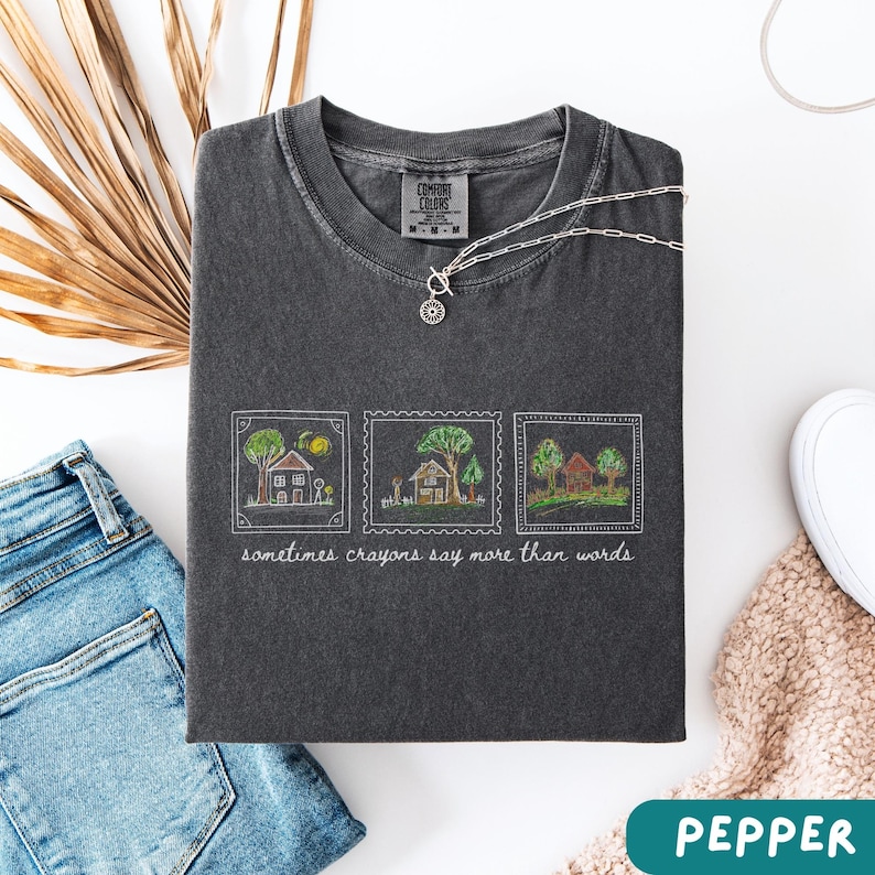 Może przedstawiać: Szara koszulka z trzema ilustracjami dom&oacute;w w ramkach i napisem "sometimes crayons say more than words". Srebrny naszyjnik z zawieszką w kształcie słońca spoczywa na koszulce. Koszulka jest złożona na białej powierzchni z dżinsami i butem.