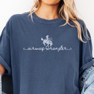 Pu&ograve; includere: Una maglietta blu con una grafica bianca di una donna a cavallo e il testo "airway wrangler" in un font corsivo.