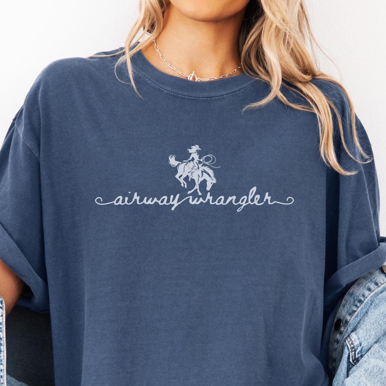 Pu&ograve; includere: Una maglietta blu con una grafica bianca di una donna a cavallo e il testo "airway wrangler" in un font corsivo.