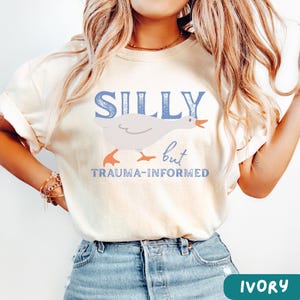 May include: Ivory-colored t-shirt with the words "SILLY but TRAUMA-INFORMED" in blue. A cartoon goose is featured in the center. The shirt is a light cream color.