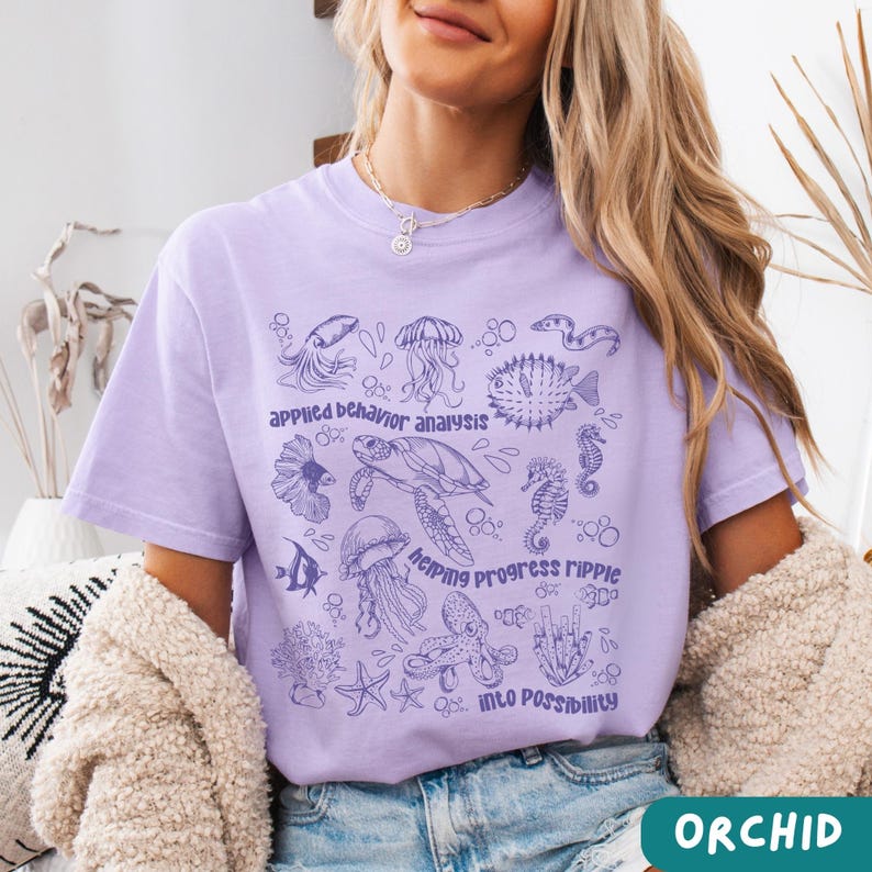 May include: A light purple t-shirt with a marine life graphic. The design includes illustrations of various sea creatures such as jellyfish, seahorses, and a turtle. Text on the shirt reads "Applied Behavior Analysis, Helping Progress Ripple Into Possibility."