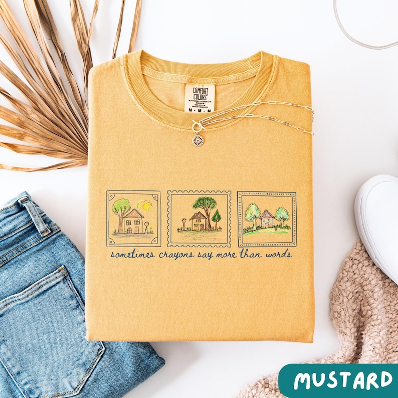 Może przedstawiać: Musztardowa bluza Comfort Colors z okrągłym dekoltem. Z przodu znajdują się trzy ilustracje dom&oacute;w i drzew w stylu znaczka pocztowego z napisem "sometimes crayons say more than words". Srebrny naszyjnik jest zarzucony na bluzę.