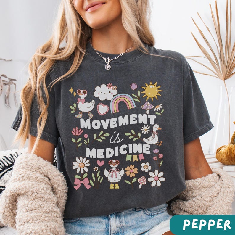 May include: A dark gray cropped t-shirt with a graphic print of a rainbow, clouds, hearts, flowers, and ducks. The text "Movement is Medicine" is printed on the shirt.
