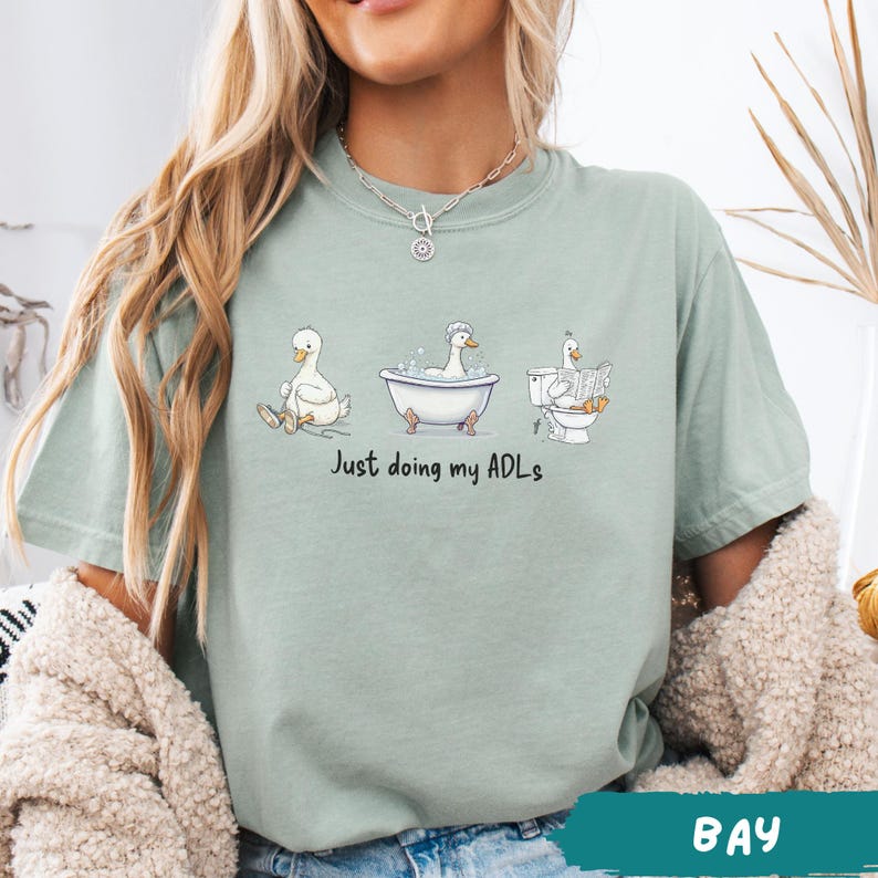 Op de afbeelding: Saliegroen T-shirt met een humoristische afbeelding van drie eenden die hun dagelijkse taken uitvoeren. De eenden worden afgebeeld terwijl ze zitten, baden en lezen op het toilet. De tekst "Just doing my ADLs" staat eronder.
