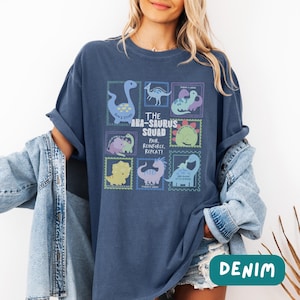 May include: A dark blue t-shirt featuring a graphic print of various cartoon dinosaurs arranged in stamp-like frames. The central text reads THE ABA-SAURUS SQUAD PAIR, REINFORCE, REPEAT! The shirt is worn with a light blue denim jacket over the shoulders.