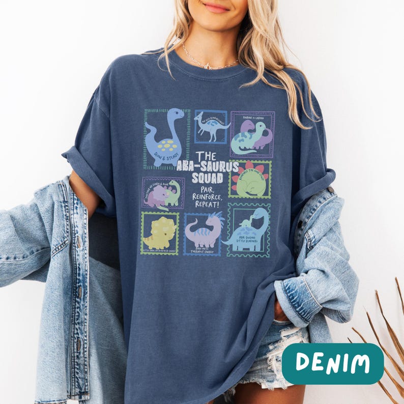 May include: A dark blue t-shirt featuring a graphic print of various cartoon dinosaurs arranged in stamp-like frames. The central text reads THE ABA-SAURUS SQUAD PAIR, REINFORCE, REPEAT! The shirt is worn with a light blue denim jacket over the shoulders.