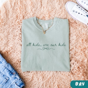 May include: A light green t-shirt with the text "all kids, are our kids" in a script font. A gold necklace rests on the shirt. The shirt is on a pink fluffy surface, with a pair of white sneakers and blue jeans visible.