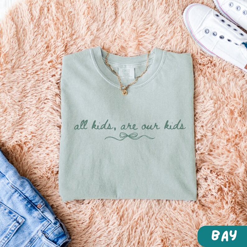 May include: A light green t-shirt with the text "all kids, are our kids" in a script font. A gold necklace rests on the shirt. The shirt is on a pink fluffy surface, with a pair of white sneakers and blue jeans visible.