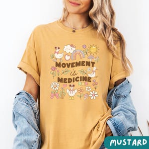 May include: A mustard yellow t-shirt with a graphic print of a rainbow, clouds, flowers, and ducks. The text "Movement is Medicine" is printed on the shirt.