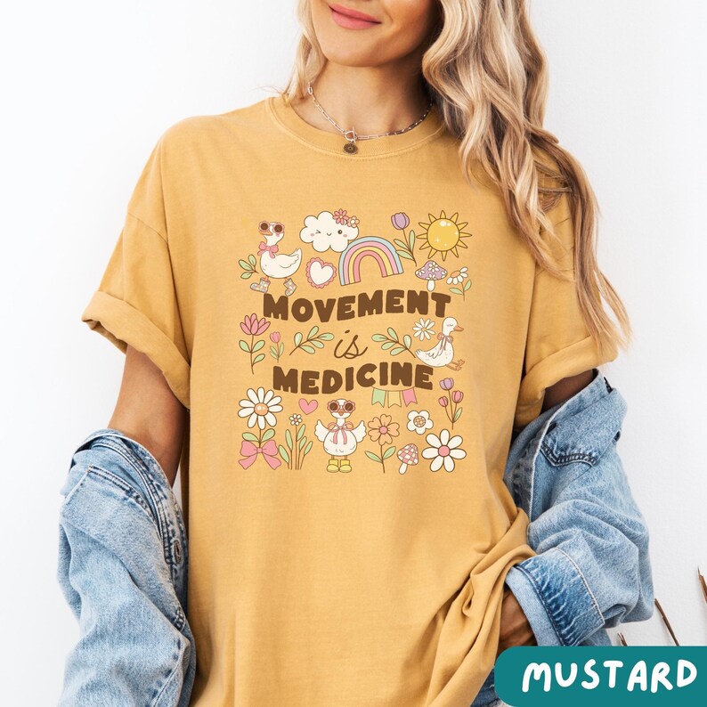 May include: A mustard yellow t-shirt with a graphic print of a rainbow, clouds, flowers, and ducks. The text "Movement is Medicine" is printed on the shirt.