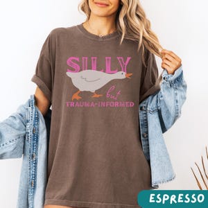 May include: Espresso-colored t-shirt with the words "SILLY" above a cartoon goose and the word "but" and "TRAUMA-INFORMED" below. The goose is white with orange feet and beak. The text is in pink.