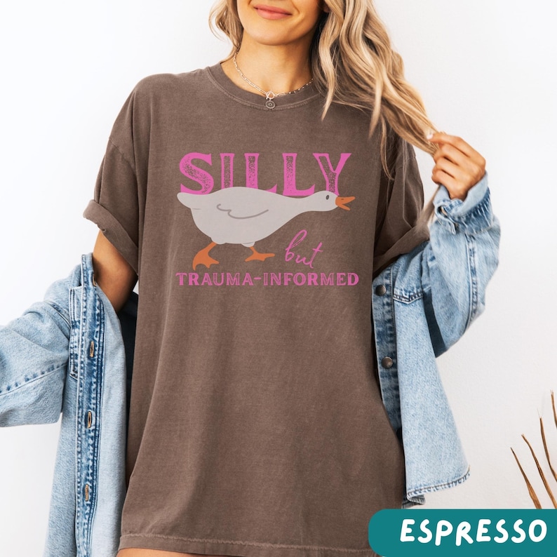 May include: Espresso-colored t-shirt with the words "SILLY" above a cartoon goose and the word "but" and "TRAUMA-INFORMED" below. The goose is white with orange feet and beak. The text is in pink.