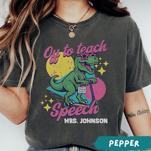 May include: A charcoal grey t-shirt featuring a green dinosaur wearing star-shaped glasses and riding a blue scooter. The graphic includes the text "Off to teach Speech" and "MRS. JOHNSON" with a "SPEECH THERAPY" sign hanging from the scooter.