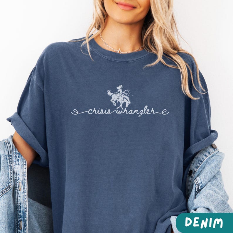 K&ouml;nnte beinhalten: Ein marineblaues T-Shirt mit einer wei&szlig;en Grafik einer Cowgirl, die ein Pferd reitet. Der Text "crisis wrangler" steht unter der Grafik.