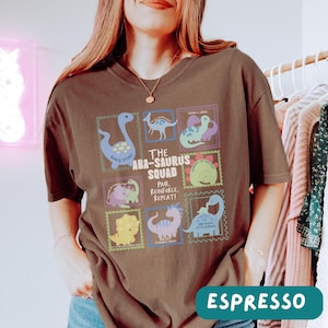 May include: A dark brown short-sleeve t-shirt featuring a graphic print of various colorful dinosaurs arranged in rectangular frames. The text reads The Aba-Saurus Squad, Pair, Reinforce, Repeat, and includes individual dinosaur names and phrases.