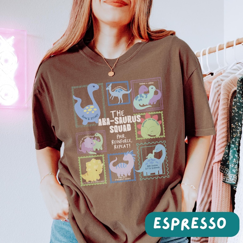May include: A dark brown short-sleeve t-shirt featuring a graphic print of various colorful dinosaurs arranged in rectangular frames. The text reads The Aba-Saurus Squad, Pair, Reinforce, Repeat, and includes individual dinosaur names and phrases.
