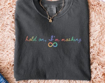 May include: Dark gray t-shirt with the text "hold on, I'm masking" in rainbow colors, along with an infinity symbol. A gold chain necklace rests on the shirt. The shirt is on a textured, light brown surface.