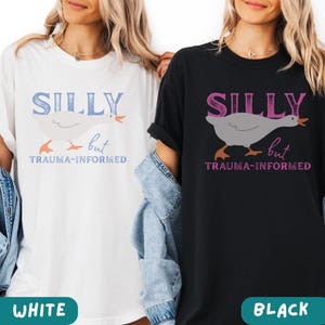 May include: Two t-shirts, one white and one black, each featuring the text "SILLY but TRAUMA-INFORMED." A cartoon goose is printed below the word "SILLY." The white shirt has blue text, and the black shirt has pink text.