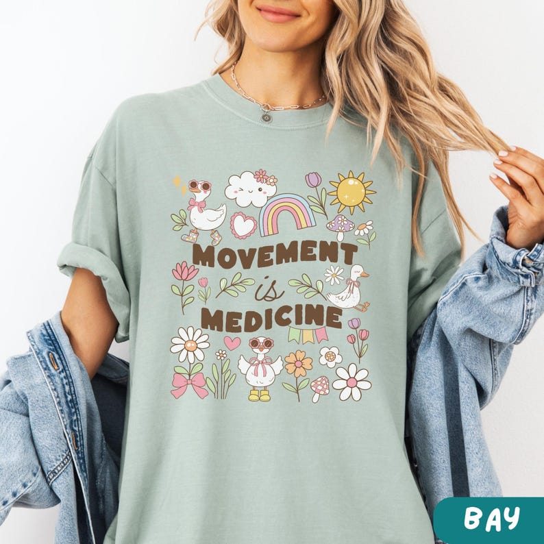 May include: A light green t-shirt with a graphic design featuring a rainbow, clouds, flowers, mushrooms, and ducks. The text "Movement is Medicine" is printed in brown on the shirt.