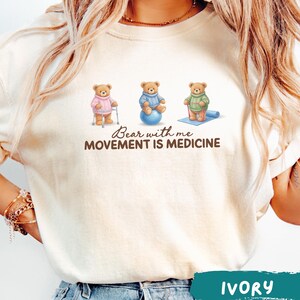 May include: Ivory-colored t-shirt featuring three cartoon bears in sweaters, each engaged in a different activity: using a walker, sitting on a blue exercise ball, and doing yoga on a mat. The text "Bear with me, Movement is Medicine" is below the bears.