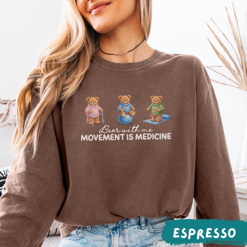 May include: Espresso-colored sweatshirt featuring three cartoon bears engaged in different activities. The bears are depicted with a cane, exercise ball, and yoga mat. The text "Bear with me, Movement is Medicine" is printed below the bears.