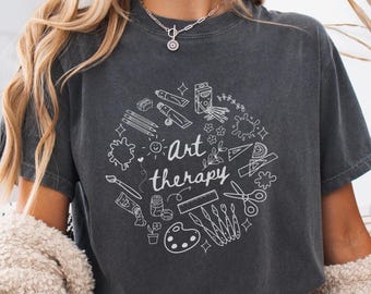 Op de afbeelding: Donkergrijs t-shirt met een wit cirkelvormig ontwerp met kunstbenodigdheden zoals verftubes, potloden en een palet. De woorden "Art therapy" zijn in een sierlijk lettertype geschreven.