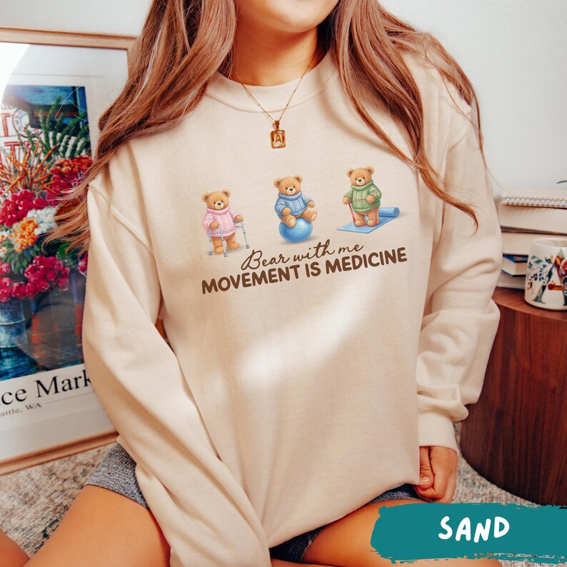 May include: Sand-colored sweatshirt featuring three cartoon bears in sweaters, with the text "Bear with me, Movement is Medicine." One bear uses a cane, another sits on a blue exercise ball, and the third stands on a yoga mat.