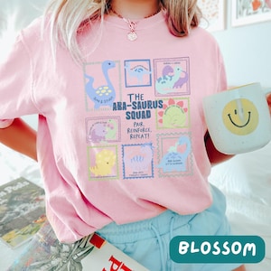 May include: A light pink t-shirt featuring a graphic print titled The ABA-Saurus Squad. The design includes multiple colorful dinosaur illustrations in rectangular frames with text, including phrases like Slow & Steady and Pair, Reinforce, Repeat.