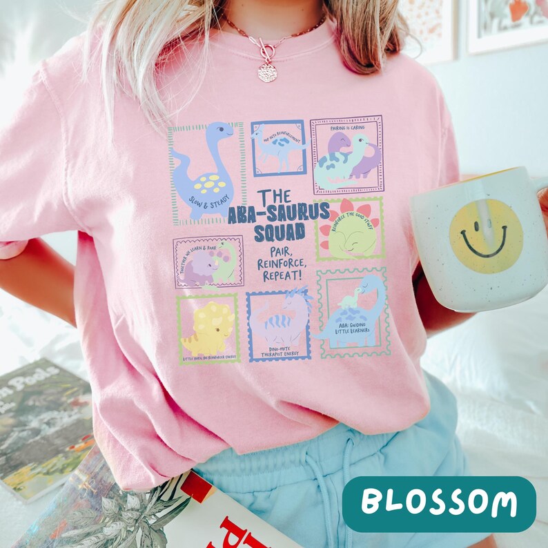 May include: A light pink t-shirt featuring a graphic print titled The ABA-Saurus Squad. The design includes multiple colorful dinosaur illustrations in rectangular frames with text, including phrases like Slow & Steady and Pair, Reinforce, Repeat.