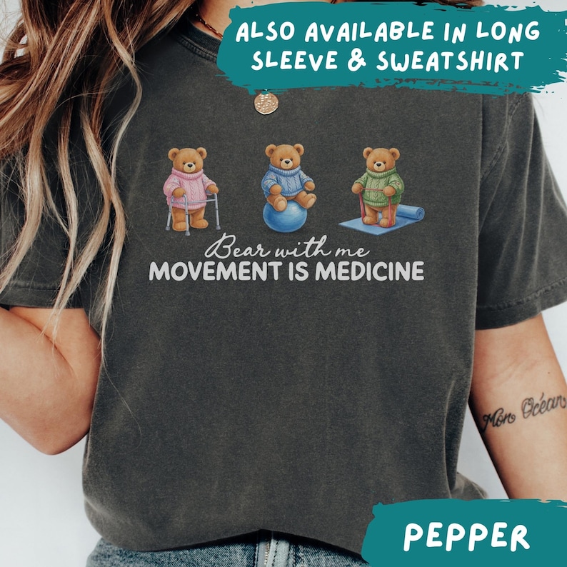 May include: Dark gray t-shirt with the text "Bear with me MOVEMENT IS MEDICINE." The shirt features three cartoon bears in sweaters, exercising with a cane, exercise ball, and yoga mat. The text "ALSO AVAILABLE IN LONG SLEEVE & SWEATSHIRT" is also visible.