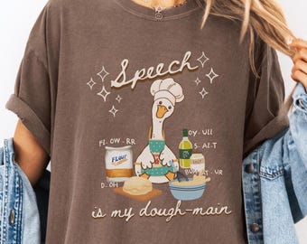 May include: Brown t-shirt with a playful graphic of a goose chef and the text "Speech is my dough-main." The design includes baking ingredients like flour, salt, and oil, and a mixing bowl.