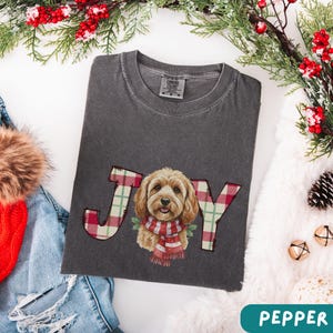 May include: Dark grey t-shirt with the word "JOY" in a plaid pattern, featuring a dog wearing a red and white scarf. The shirt is a Comfort Colors brand.