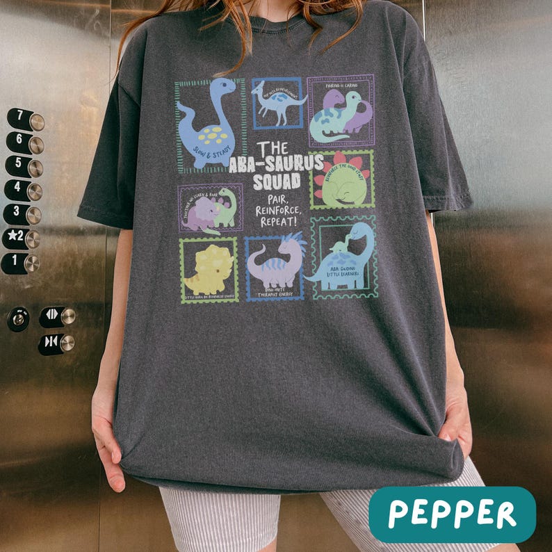 May include: A charcoal gray short-sleeve t-shirt featuring a graphic print of various colorful dinosaurs in postage stamp-style frames. The central text reads The ABA-SAURUS SQUAD, Pair, Reinforce, Repeat! The design includes light blue, green, and purple dinosaurs.