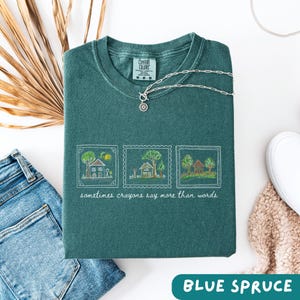 Może przedstawiać: Złożona koszulka Comfort Colors w kolorze niebieskiego świerku z białym haftowanym wzorem trzech ilustracji dom&oacute;w. Pod ilustracjami znajduje się napis "sometimes crayons say more than words". Srebrny naszyjnik leży na koszulce.