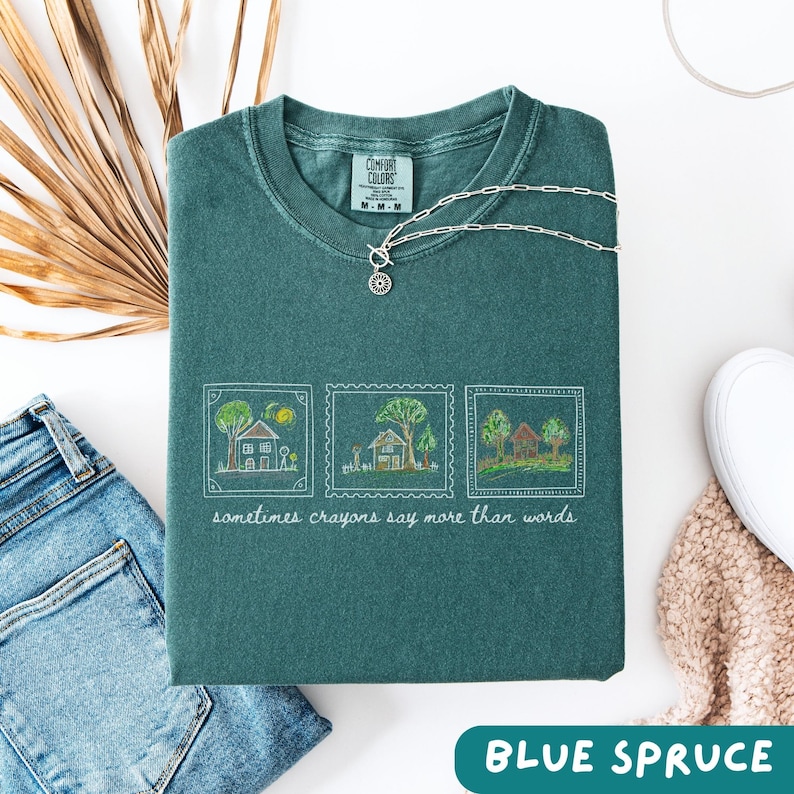 Może przedstawiać: Złożona koszulka Comfort Colors w kolorze niebieskiego świerku z białym haftowanym wzorem trzech ilustracji dom&oacute;w. Pod ilustracjami znajduje się napis "sometimes crayons say more than words". Srebrny naszyjnik leży na koszulce.