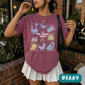 May include: A maroon short-sleeve t-shirt featuring a graphic print of various dinosaurs in colorful frames with the text THE ABA-SAURUS SQUAD PAIR, REINFORCE, REPEAT! paired with a white pleated skirt and a black shoulder bag.