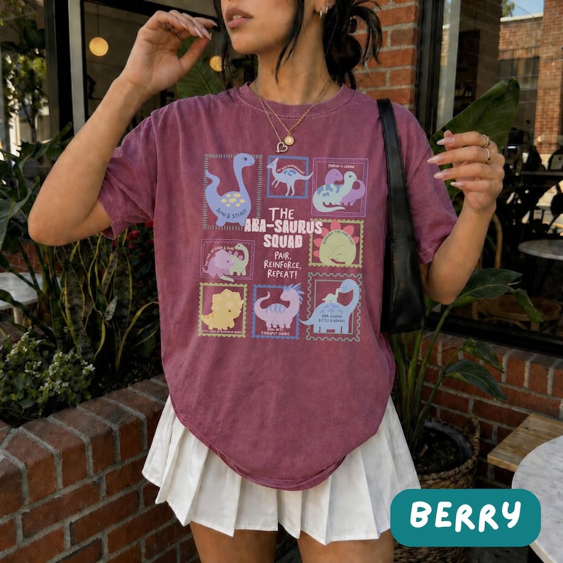 May include: A maroon short-sleeve t-shirt featuring a graphic print of various dinosaurs in colorful frames with the text THE ABA-SAURUS SQUAD PAIR, REINFORCE, REPEAT! paired with a white pleated skirt and a black shoulder bag.