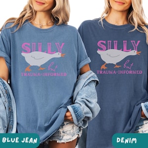 May include: Two t-shirts, one blue jean and one denim, with the text "SILLY but TRAUMA-INFORMED" in pink. Each shirt features a cartoon goose graphic. The shirts are short-sleeved and have a relaxed fit.
