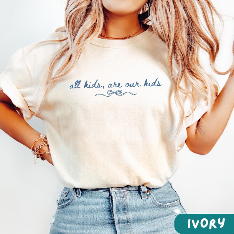 May include: Ivory-colored t-shirt with the text "all kids, are our kids" in a dark blue script. A decorative bow design is below the text. The shirt is a light cream color and has short sleeves.
