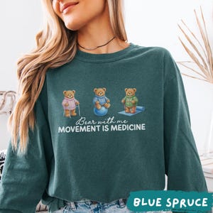 May include: Blue Spruce sweatshirt featuring three teddy bears in sweaters, each engaged in a different activity. The text "Bear with me, Movement is Medicine" is printed below the bears. The words "BLUE SPRUCE" are printed at the bottom.