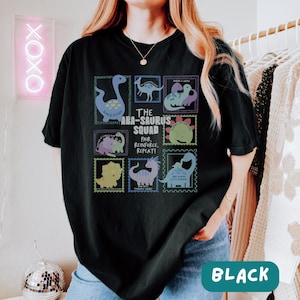 May include: A black short-sleeve t-shirt featuring a graphic print of various dinosaurs in colorful postage stamp-style frames. The central text reads: THE ABA-SAURUS SQUAD, PAIR, REINFORCE, REPEAT! Each dinosaur illustration is accompanied by small text.