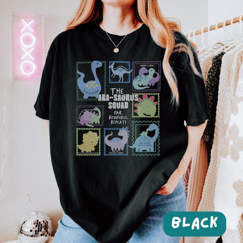 May include: A black short-sleeve t-shirt featuring a graphic print of various dinosaurs in colorful postage stamp-style frames. The central text reads: THE ABA-SAURUS SQUAD, PAIR, REINFORCE, REPEAT! Each dinosaur illustration is accompanied by small text.